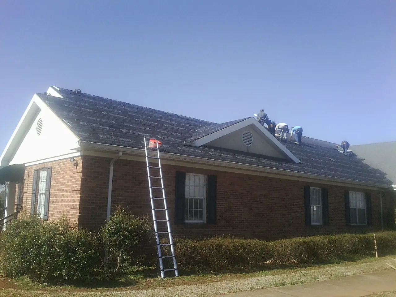 Crew tearing shingles off a brick ranch roof with a ladder set — Metal Roof Repair work by our crew in Summerfield